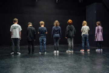 viele Kinder-Schauspieler:innen stehen mit dem Rücken zum Publikum, dahinter ist eine schwarze Wand